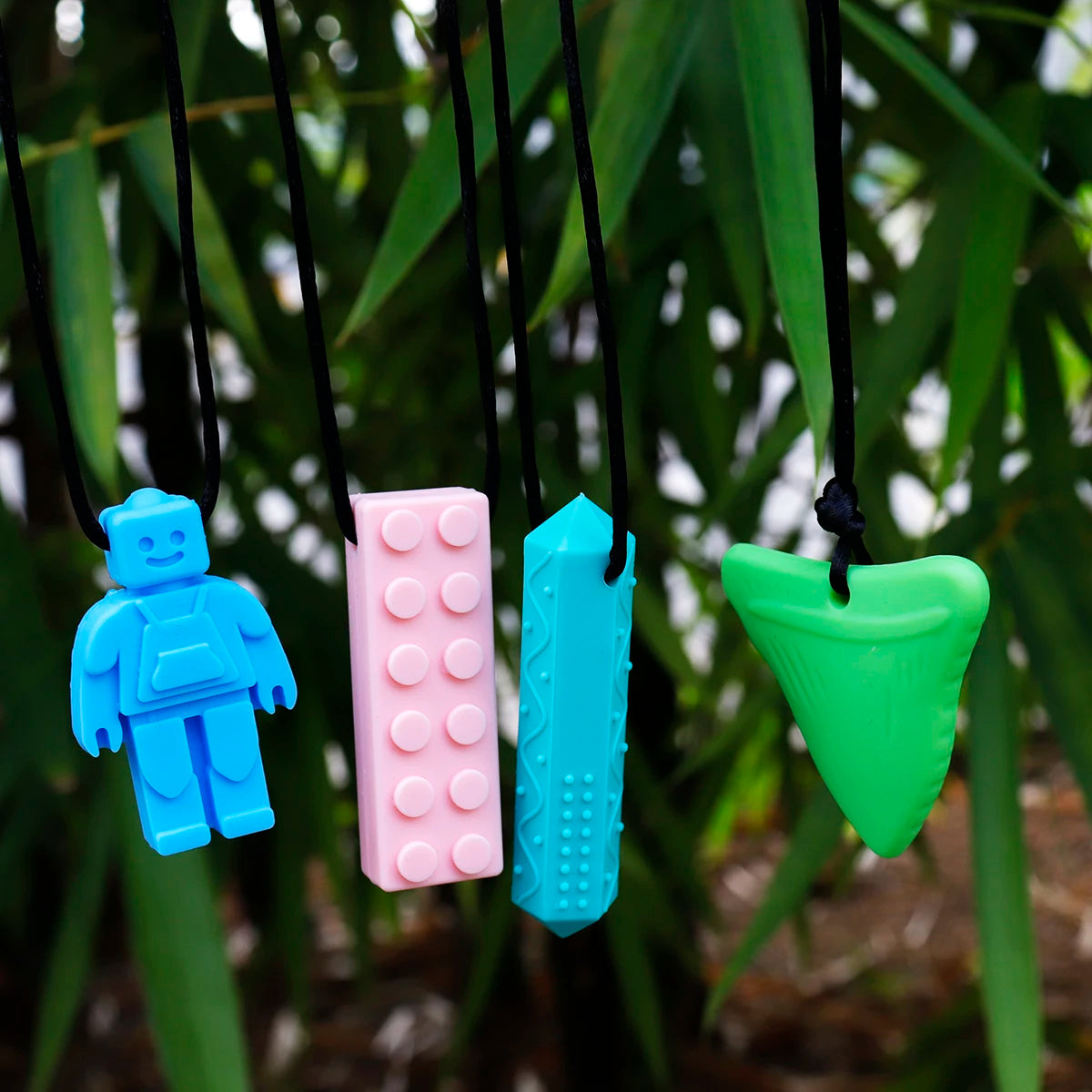 Four colorful silicone keychains shaped like a robot, block, shark tooth, and another design hanging against a green leafy background.